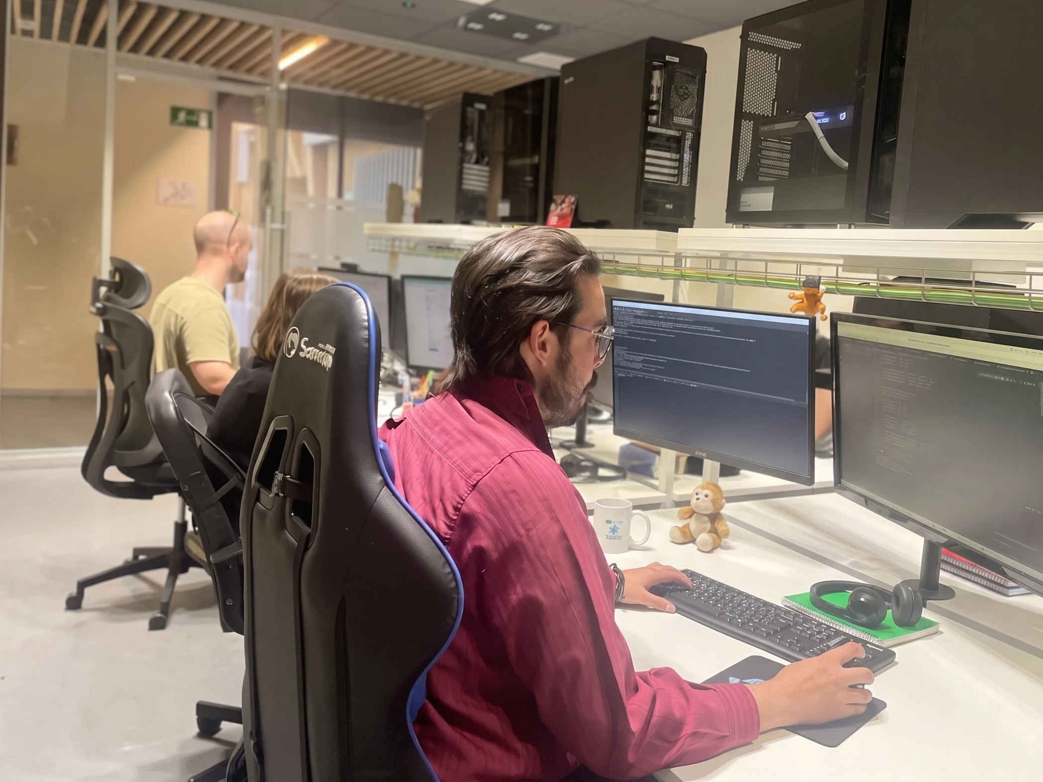 Hombre con camisa granate y gafas programando frente a dos monitores en un estudio u oficina rodeado de compañeros.